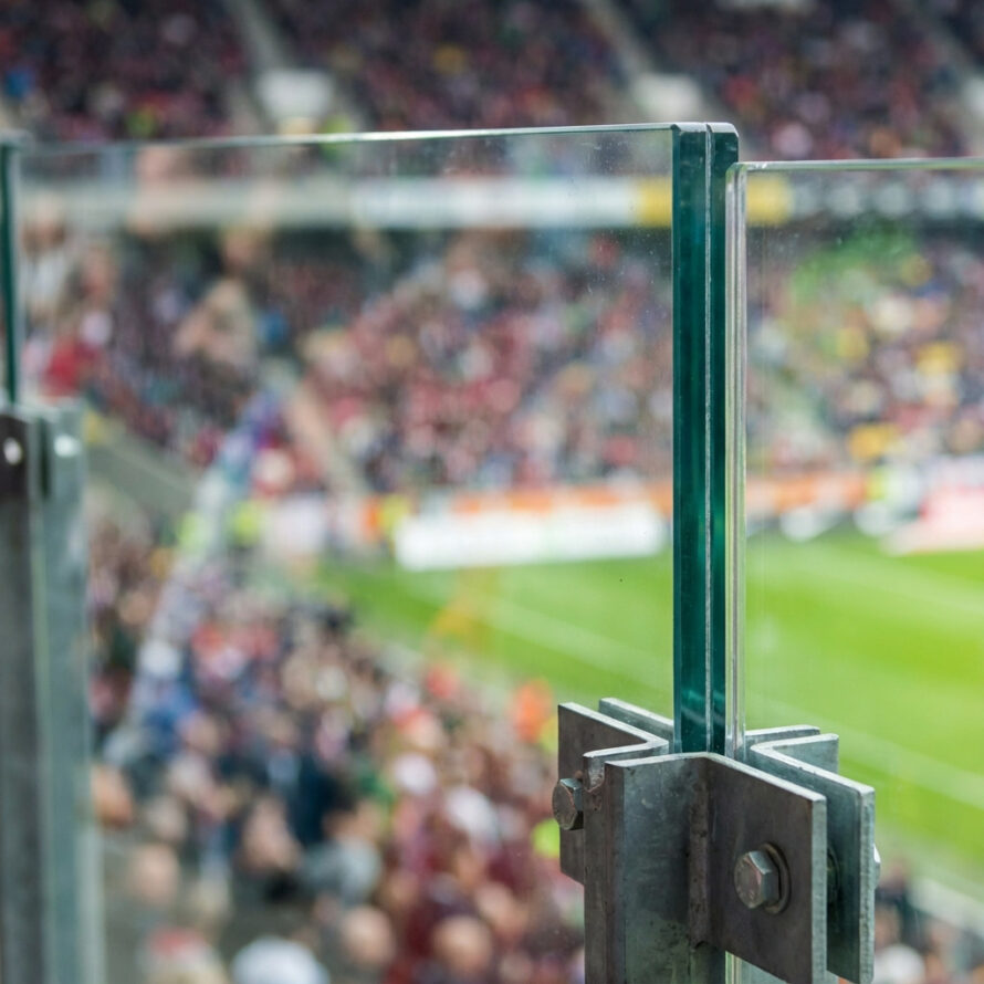 Detail van een polycarbonaat stadionwand met metalen klemconstructie, met het voetbalveld en publiek op de achtergrond.