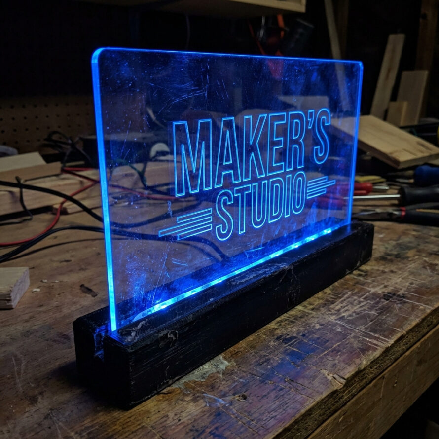 Edge-lit plexiglas LED lamp met blauw verlichte gravure “Make More Stuff” op zwarte voet.