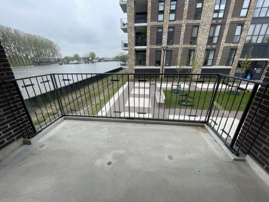 Balkon met zwarte metalen balustrade met verticale spijlen, uitzicht op een gracht en appartementencomplex.