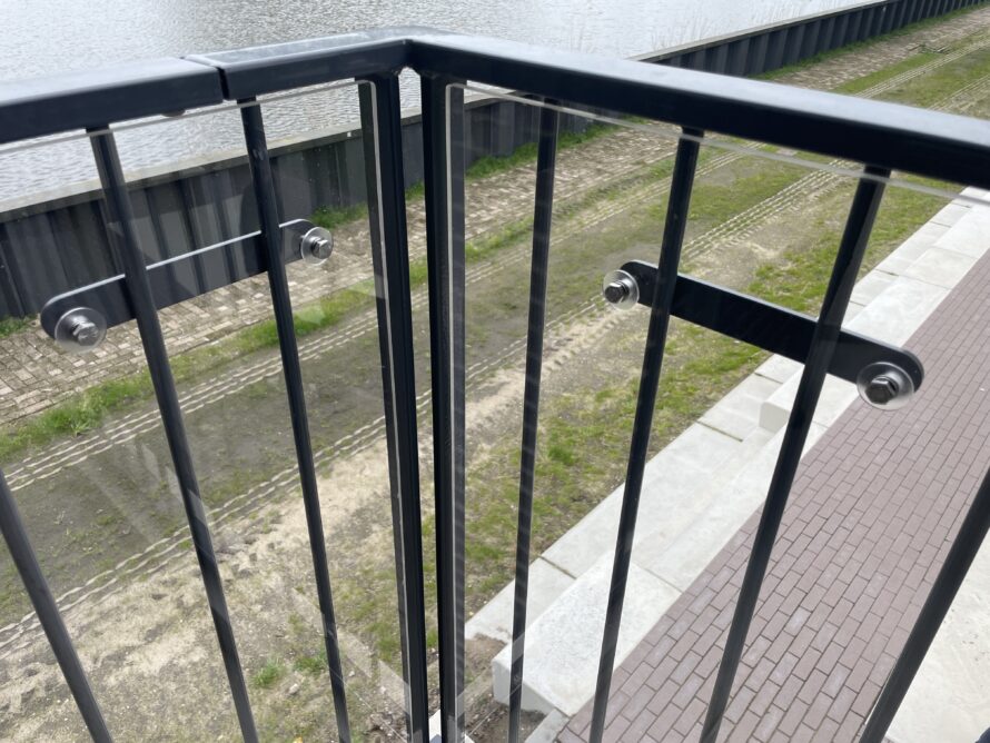 Detail van een zwarte metalen balustrade met verticale spijlen en een glasplaat bevestigd met klemmen.