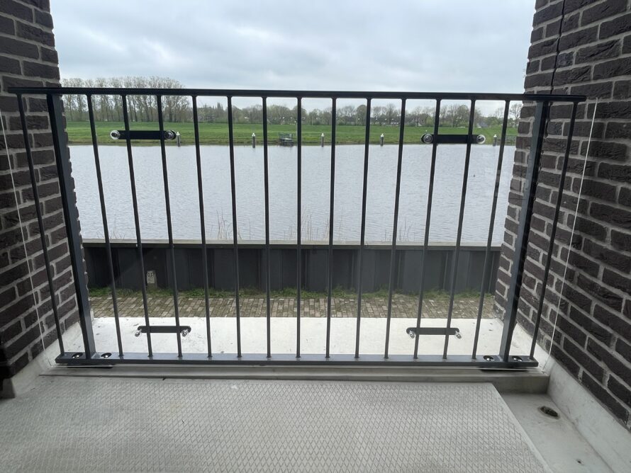 Balkon met zwarte metalen balustrade met verticale spijlen tussen twee bakstenen muren, met uitzicht op water en groen.