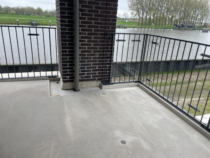 Leeg balkon met grijze vloer, metalen railing en een bakstenen muur, met uitzicht op water en groen landschap.