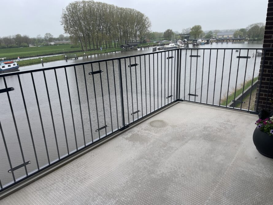 Balkon met grijze betonnen vloer en zwarte metalen railing, met uitzicht op een meer en bomen op de achtergrond.