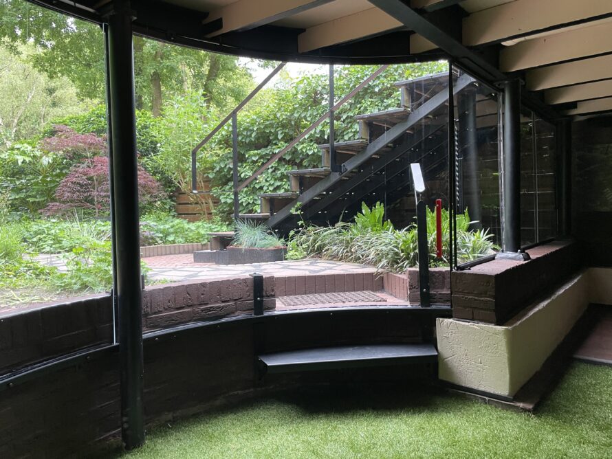 Binnenzijde van een ronde plexiglas serre met zitbank en uitzicht op de tuin.