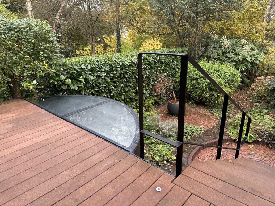 Toegang tot een gebogen plexiglas serre vanaf een houten terras met glazen railing.