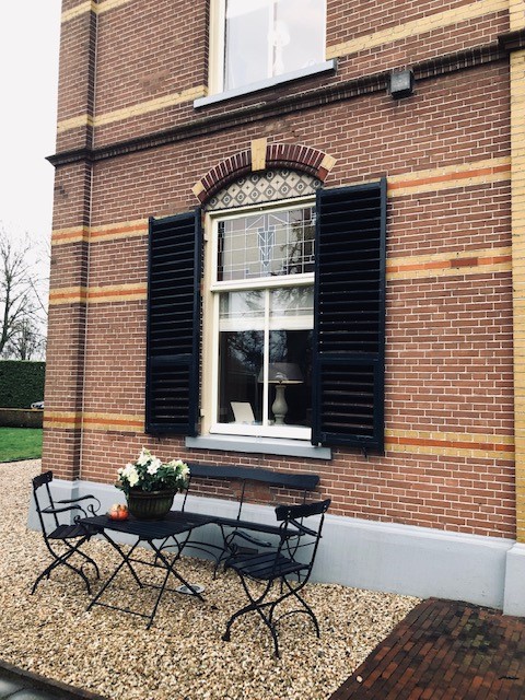 Raam met zwarte luiken bij een terras met kleine tuintafel en stoelen.